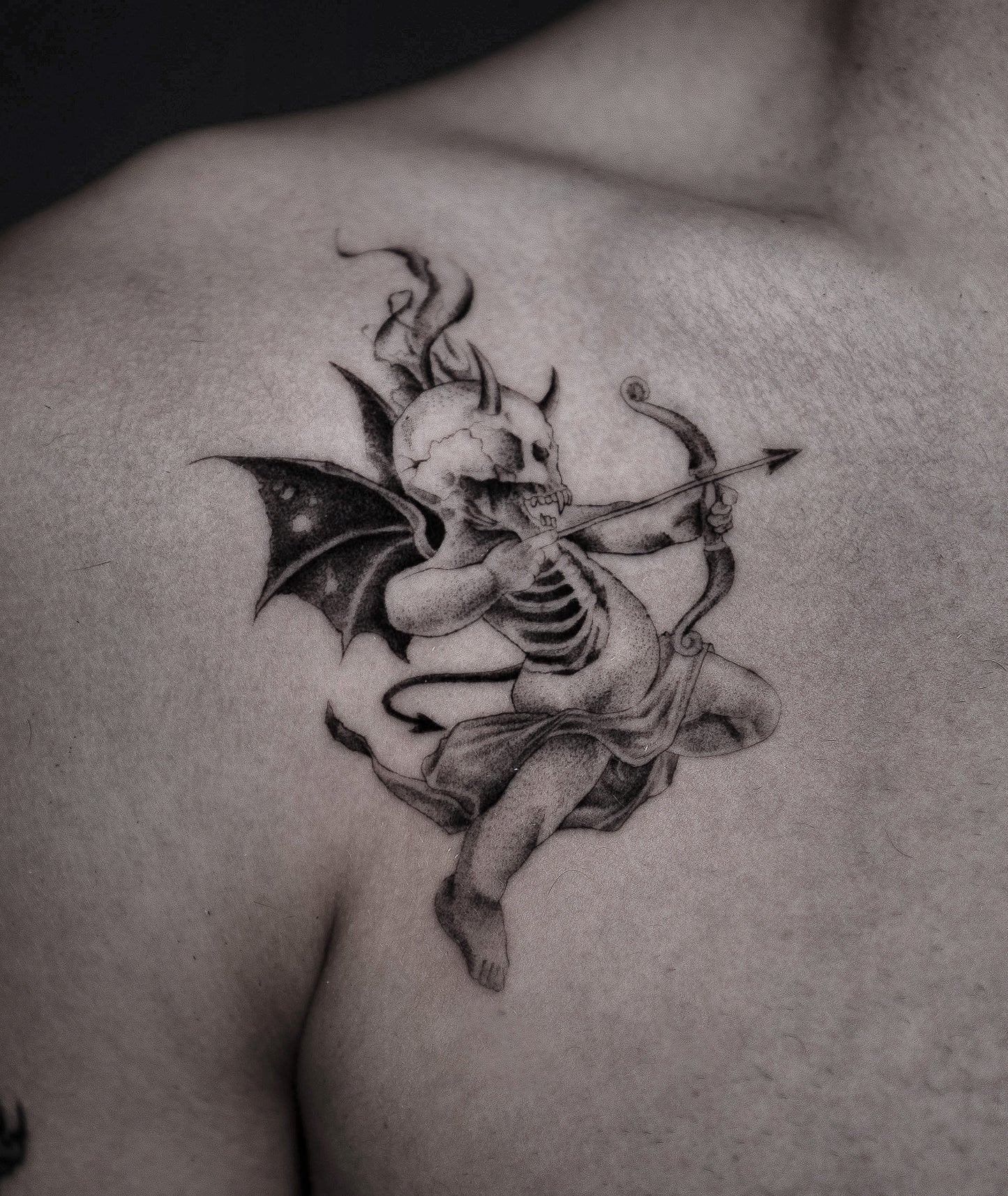 A detailed black and gray tattoo of a skeletal cupid with bat wings playing a bow on a person's upper shoulder.