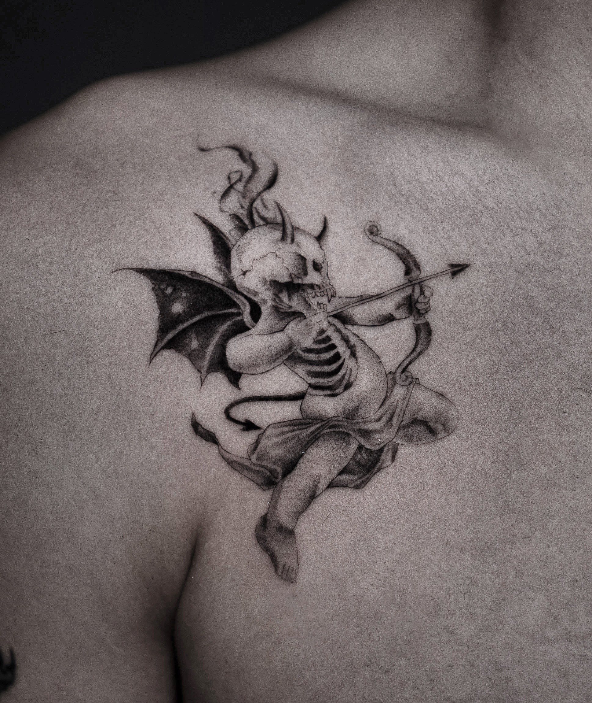 A detailed black and gray tattoo of a skeletal cupid with bat wings playing a bow on a person's upper shoulder.